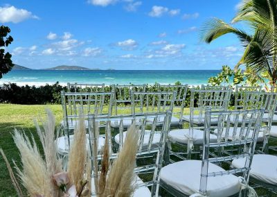 Beachfront bvi wedding venue setup in the British Virgin Islands with clear chairs, floral arrangements, and turquoise ocean views under a sunny sky.