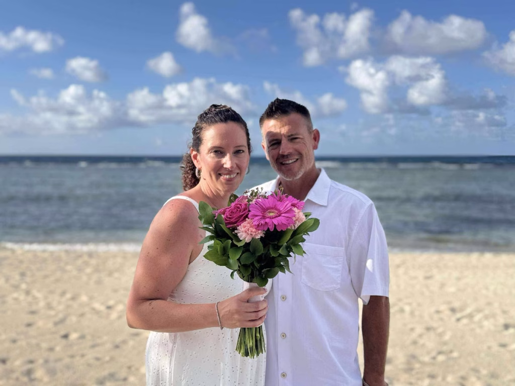 Couple on the beach at Scrub Island Resort in the British Virgin Islands, holding a bouquet of pink flowers with the ocean in the background.